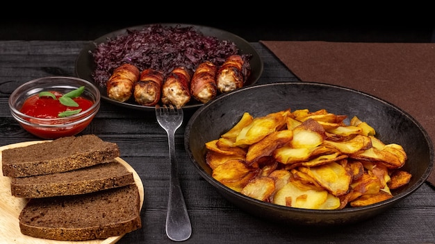 Pieczony ziemniak w garnku, kawałki czarnego chleba żytniego na drewnianym talerzu do serwowania, domowe kiełbaski w boczku i duszona kapusta na czarnym rustykalnym tle.