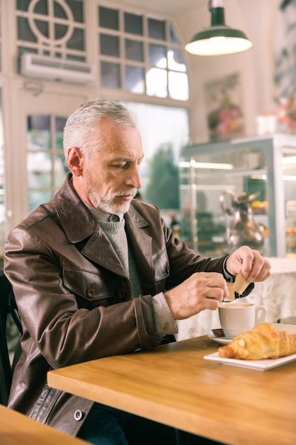 Piekarnia francuska. Dojrzały brodaty siwy mężczyzna ubrany w brązową skórzaną kurtkę siedzi we francuskiej piekarni