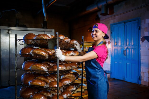 Piekarz dziewczyna bierze gorący chleb w piekarni na tle regałów z chlebem. Przemysłowa produkcja chleba. Etap pieczenia w piekarni