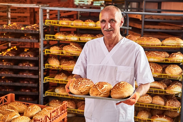 Piekarz trzyma w rękach tacę ze świeżym gorącym chlebem na tle półek z wypiekami w piekarni Przemysłowa produkcja chleba