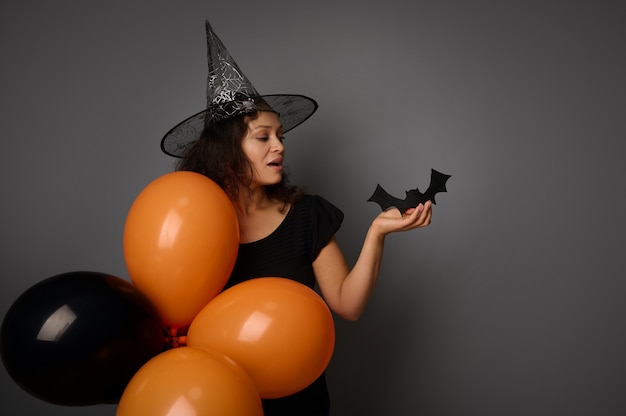 Piękna Afroamerykanka w kapeluszu czarodzieja, ubrana na czarno, trzyma pomarańczowe balony i ręcznie wycięty czarny nietoperz, pozuje na szarym tle z miejscem na reklamę. Koncepcja przyjęcia na Halloween
