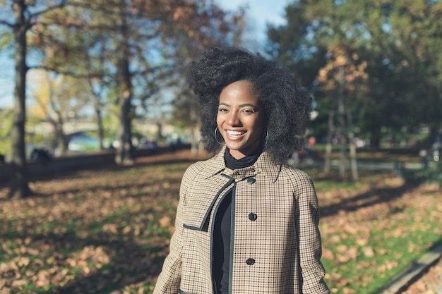 Piękna afroamerykańska młoda kobieta z fryzurą afro w stylowym płaszczu w parku uśmiechnięta koncepcja sezonu jesiennego i stylu życia