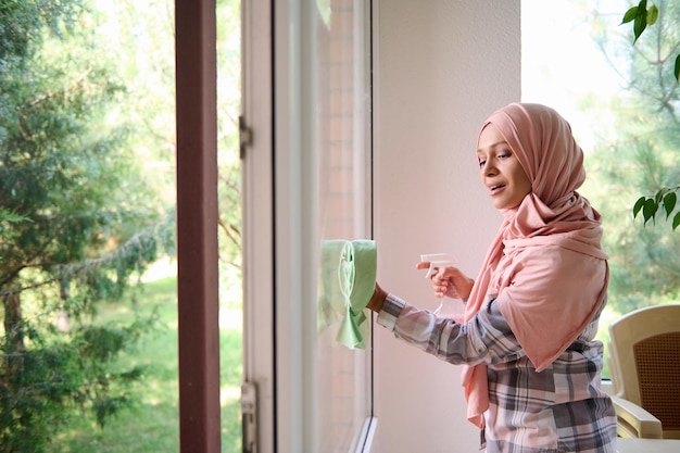 Piękna Arabska Muzułmanka z zakrytą głową w hidżabie myje okna, spryskuje detergentem i wyciera gąbką, ciesząc się wiosennym porządkiem w domu