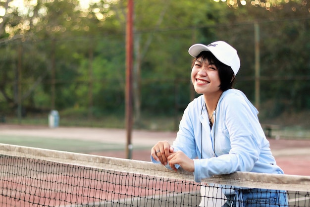 Zdjęcie piękna azjatka z krótkimi włosami, w kapeluszu i szeroko uśmiechnięta na korcie tenisowym