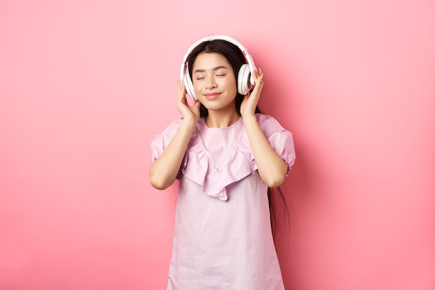 Piękna Azjatka zamyka oczy słuchając muzyki w słuchawkach, ciesząc się cichym dźwiękiem, stojąc na różowym tle.