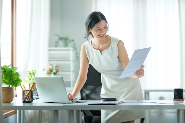 Piękna azjatycka bizneswoman korzystająca z laptopa i sprawdzająca dokument finansowy przy biurku