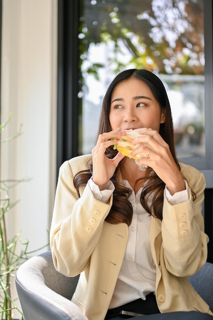 Piękna azjatycka bizneswoman siedzi na zewnątrz w kawiarni jedząc kanapkę