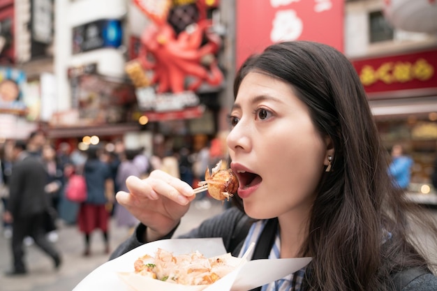 piękna azjatycka kobieta jedzenie takoyaki. biorąc kęs orientalnego jedzenia. widok z boku