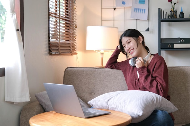 Piękna Azjatycka kobieta korzysta z laptopa podczas słuchania słuchawek siedząc na kanapie w domu