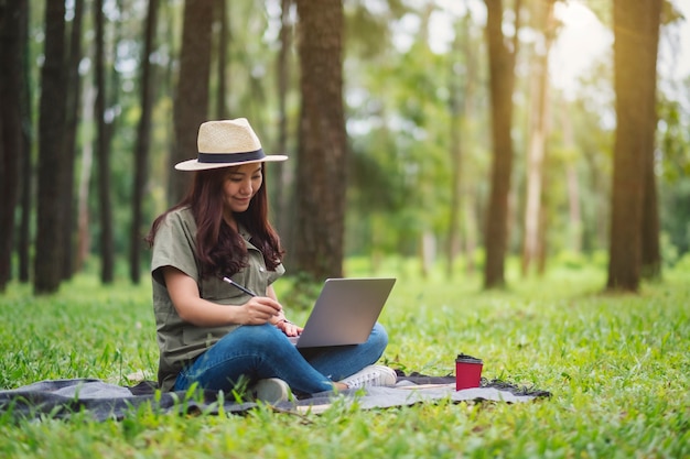 Piękna azjatycka kobieta pracująca i pisząca na klawiaturze laptopa siedząc w parku