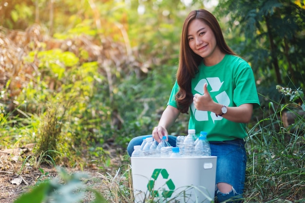 Piękna azjatycka kobieta robiąca kciuk w górę znak ręką podczas zbierania plastikowych butelek na śmieci do kosza na zewnątrz