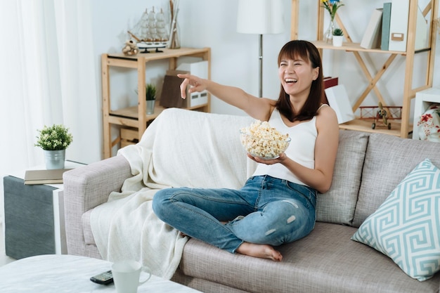 piękna azjatycka kobieta siedząca z popcornem na kanapie ogląda zabawny pokaz w swoim miejscu. Chinka śmieje się i wskazuje na ekran, żeby zrobić kilka śmiesznych dowcipów, które opowiada gospodarz telewizji.