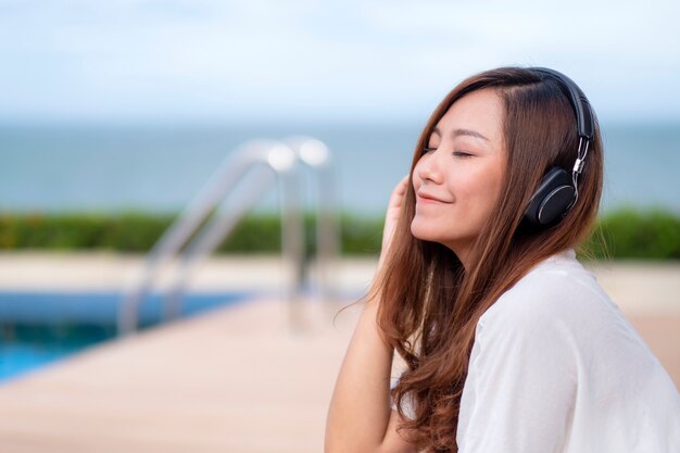 Piękna azjatycka kobieta słuchająca muzyki przez słuchawki siedząc przy basenie