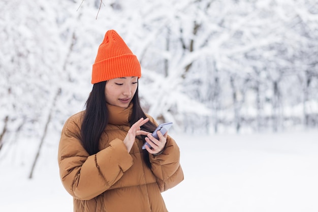 Piękna azjatycka kobieta spacerująca po parku używa telefonu do zakupów online w zimowy, śnieżny dzień