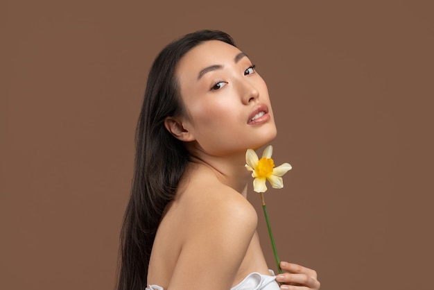 Piękna azjatycka kobieta trzymająca piękny kwiat żonkila i patrząca na kamerę pozującą na brązowym studio