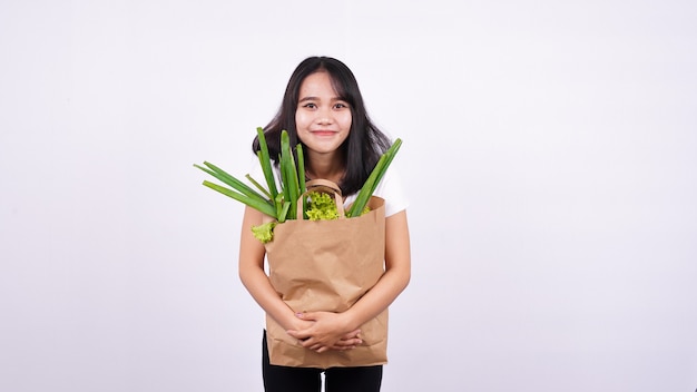Piękna azjatycka kobieta uśmiecha się z papierową torbą świeżych warzyw z izolowaną białą powierzchnią