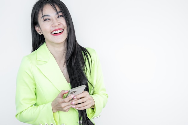 Piękna Azjatycka kobieta w zielonym garniturze pastel trzyma inteligentny telefon i uśmiecha się na białym tle Szczęśliwa radosna dziewczyna z świętowaniem zwycięstwa Azjatycka kobieta udanego szczęścia