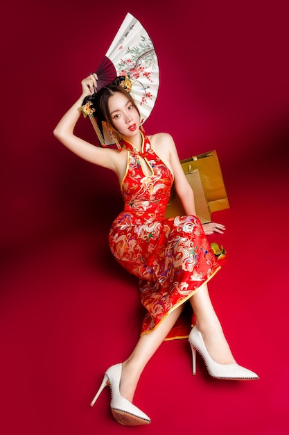 Piękna azjatycka kobieta z czystą świeżą skórą ubrana w sukienkę cheongsam trzymająca wentylator siedzący ze złotymi torbami na zakupy na czerwonym tle Portret modelki w studio Szczęśliwego chińskiego nowego roku