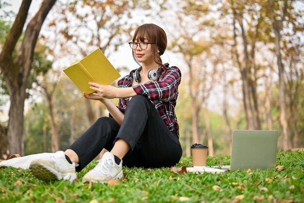 Piękna azjatycka studentka college'u siedzi na trawie i czyta książkę