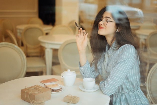 Piękna azjatykcia dziewczyna cieszy się gorącą herbaty w kawiarni podczas gdy słuchający muzykę na hełmofonach, widok przez nadokiennego szkła