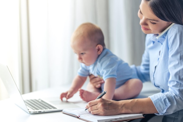 Piękna Biznesowa Dama W Klasycznym Garniturze Rozmawia Przez Telefon Komórkowy, Robiąc Notatki I Uśmiechając Się, Pracując W Biurze, Podczas Gdy Jej Słodkie Małe Dziecko Siedzi Na Biurku