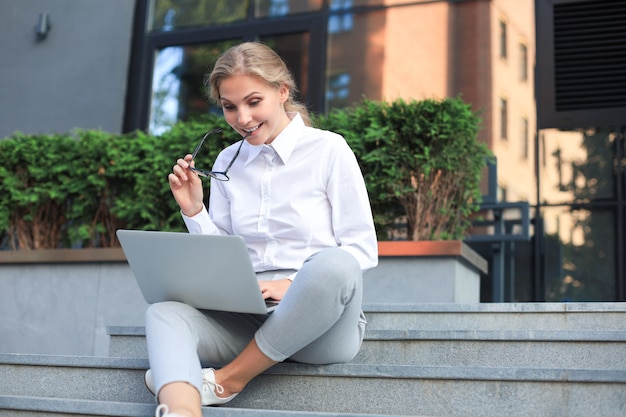 Piękna biznesowa kobieta siedzi w pobliżu centrum biznesowego i za pomocą laptopa.