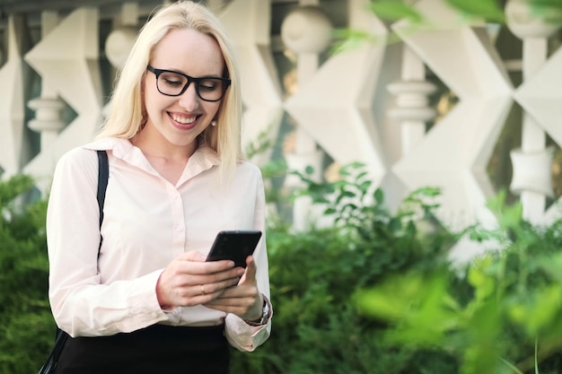 Piękna biznesowa kobieta w czarnej koszuli i okularach w parku z zielonymi krzakami trzyma telefon komórkowy w dłoniach raduje się i uśmiecha Spędza wolny czas w mediach społecznościowych