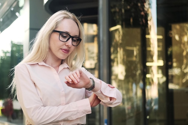 Piękna biznesowa kobieta w koszulowej czarnej spódnicy i okularach na tle wejścia do centrum biznesowego Podnosi rękę patrzy na zegarek i wychodzi z kadru