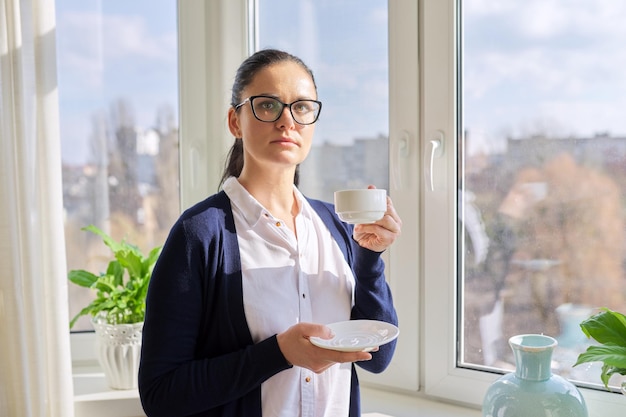 Piękna biznesowa kobieta w okularach z filiżanką kawy w pobliżu okna