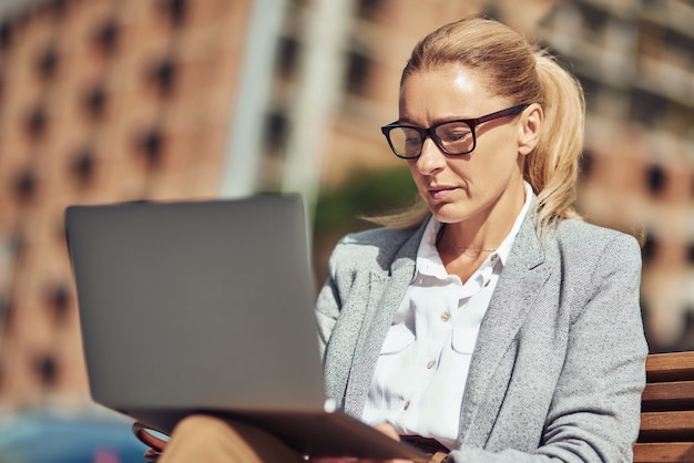 Zdjęcie piękna biznesowa kobieta w średnim wieku pracująca online przy użyciu laptopa siedząc na ławce