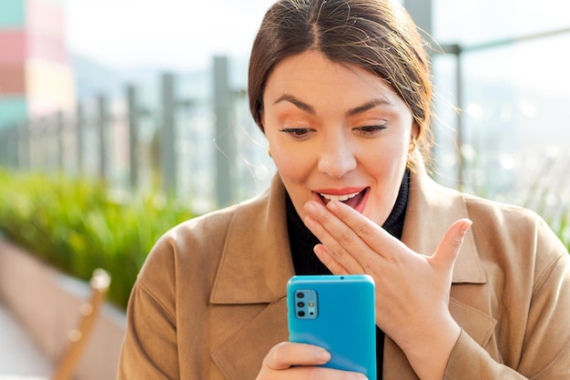 Zdjęcie piękna bizneswoman patrzy na telefon komórkowy z niespodziewaną dłonią zakrywającą usta na znak zdziwienia wiadomościami, które otrzymuje