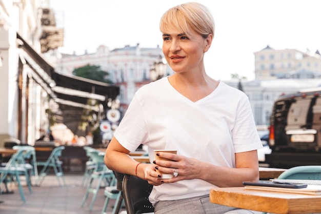 piękna blond dorosła kobieta ubrana w białą koszulkę siedzi w ulicznej kawiarni latem i pije kawę z papierowego kubka