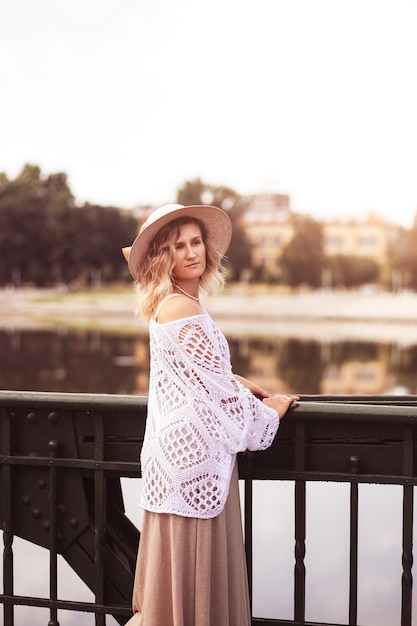 Piękna blond młoda kobieta ubrana w sukienkę i słomkowy kapelusz, ciesząc się wschodem słońca nad brzegiem morza w starym europejskim mieście Moda i styl Letnie podróże