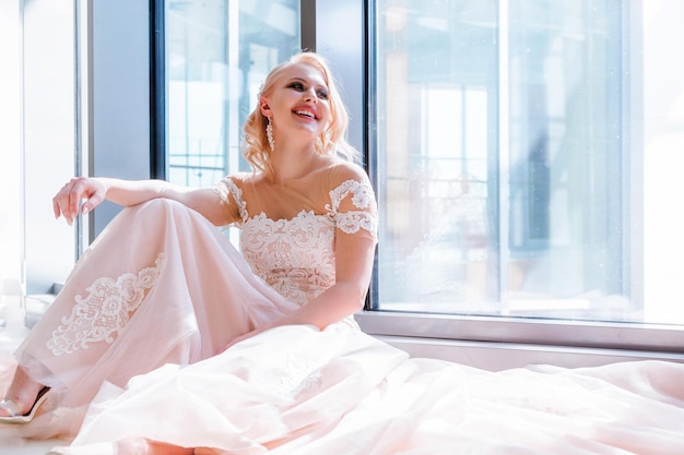 Piękna blond panna młoda w luksusowej sukni ślubnej siedzi blisko dużego okna Słońce świeci na dziewczynę rąbek tańca weselnego leży na podłodze