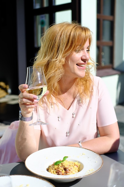 Piękna blondynka dorosła kobieta siedzi przy stoliku w kawiarni pije białe wino je spaghetti i cieszy się pogodą Kobieta uśmiecha się i patrzy w kamerę