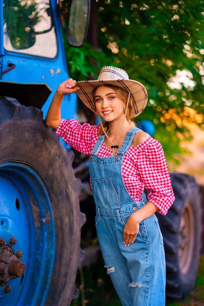 Zdjęcie piękna blondynka kowbojka w słomkowym kapeluszu i drelichu ogólnie stojąca w pobliżu niebieskiego ciągnika.