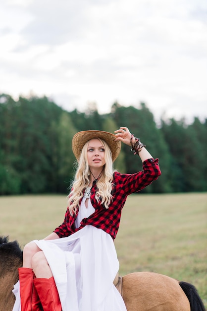 Zdjęcie piękna blondynka młoda kobieta w eleganckim kapeluszu w pobliżu starego drewnianego ogrodzenia z panną młodą kucyków