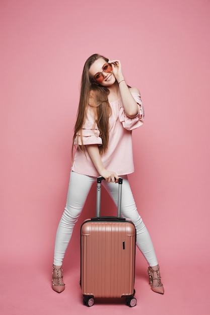 Piękna blondynka model dziewczyna w modnej bluzce z nagimi ramionami i stylowe okulary stoi z walizką i pozowanie na różowym tle w studio
