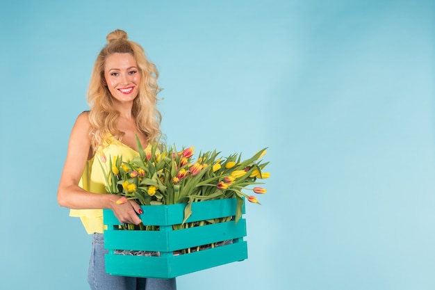 Piękna blondynka ogrodnik kobieta trzyma pudełko z tulipanami na niebieskiej ścianie z miejsca na kopię