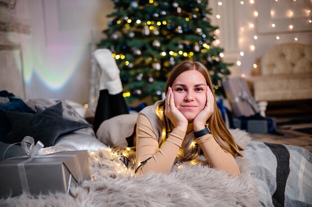Piękna blondynka siedzi przy choince w noworocznej atmosferze i raduje się Kobieta jest w jasnej, wygodnej sypialni pięknie udekorowanej na Boże Narodzenie