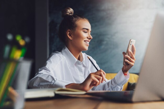 Piękna blondynka siedzi przy stole przed laptopem i trzyma w ręku smartfon.