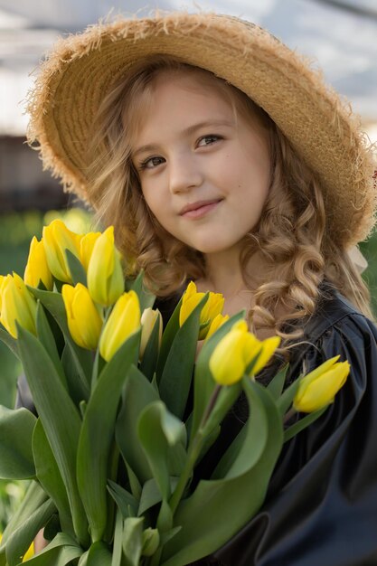piękna blondynka w czarnej sukience i słomkowym kapeluszu trzyma w dłoniach żółte wiosenne tulipany