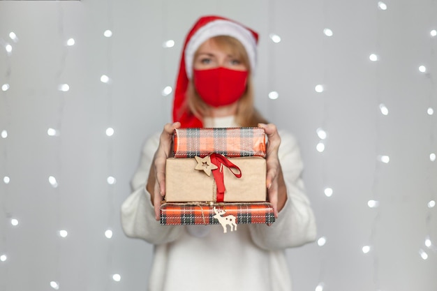 Piękna blondynka w czerwonej masce ochronnej czapki Mikołaja i białym swetrze trzyma pudełka z prezentami