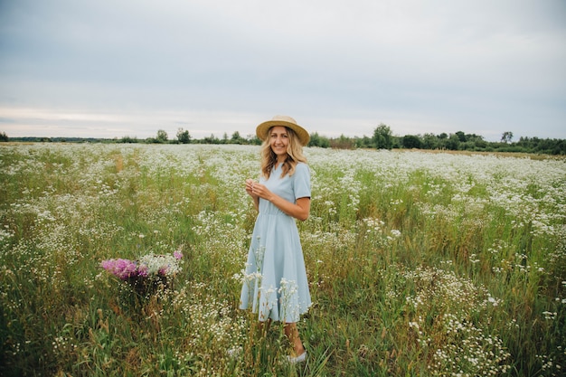 piękna blondynka w polu stokrotki. kobieta w niebieskiej sukience w polu białych kwiatów. dziewczyna z bukietem stokrotek. letnie zdjęcie przetargowe we wsi. kwiaty. dziewczyna w słomkowym kapeluszu