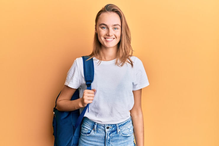 Zdjęcie piękna blondynka w studentskim plecaku wygląda pozytywnie i szczęśliwie, stoi i uśmiecha się z pewnym siebie uśmiechem pokazującym zęby.