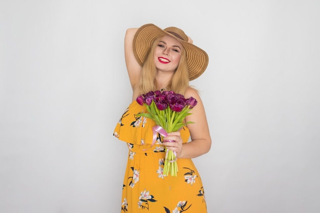 Piękna blondynka w żółtej sukience kwiatowy i słomkowy kapelusz trzymając bukiet fioletowych tulipanów