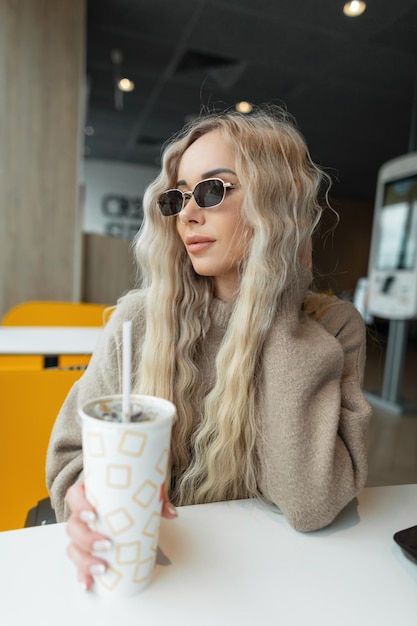 Piękna blondynka z kędzierzawą fryzurą w modnych ubraniach z okularami przeciwsłonecznymi siedzi w kawiarni i trzyma papierową filiżankę coli Kobieta pije napój w pomieszczeniu