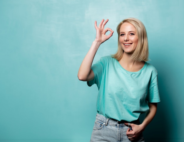 Piękna blondynki kobiety przedstawienia OK gest na błękit ścianie