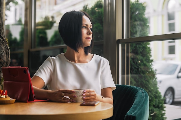 piękna brunetka biznesowa kobieta siedzi w kawiarni, pije kawę i wygląda przez okno