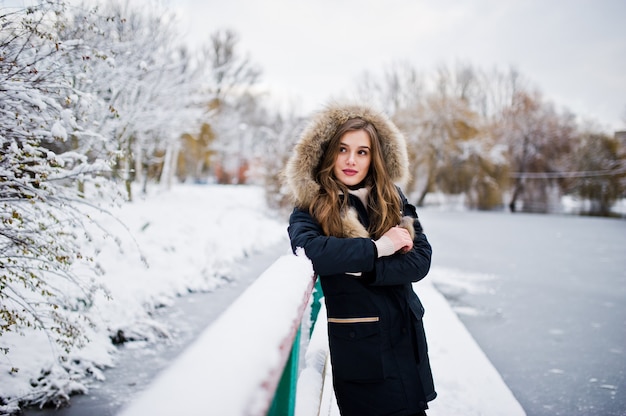 Piękna brunetka dziewczyna w zimowej ciepłej odzieży. Model na zimowej kurtce przed zamarzniętym jeziorem w parku.